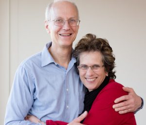Tall smiling white man in pale blue shirt hugging shorter, dark-haired white woman in red and black sweater. Both wear glasses and smile broadly. Photo by Kelly Fetcher Photography